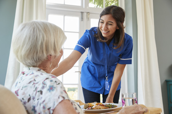 care-nurse-serving-dinner-to-a-senior-woman-at-hom-PQHJS4A